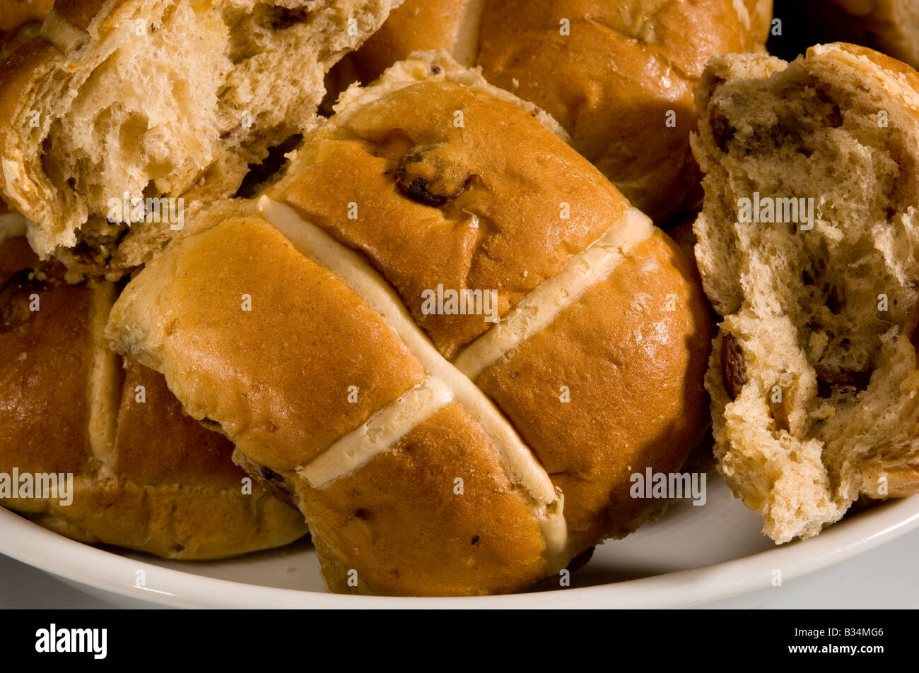 Closeup of commercially baked Hot cross buns on a white plate with one of the buns torn open to expose the dried fruit inside. UK Stock Photo