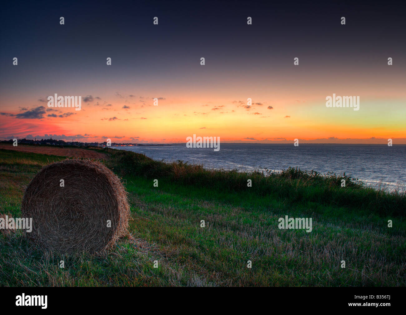 Taken in the country near Grancamp Maisy, Normandy, is a sunset scene of a field with a hay bale and the ocean in the background Stock Photo