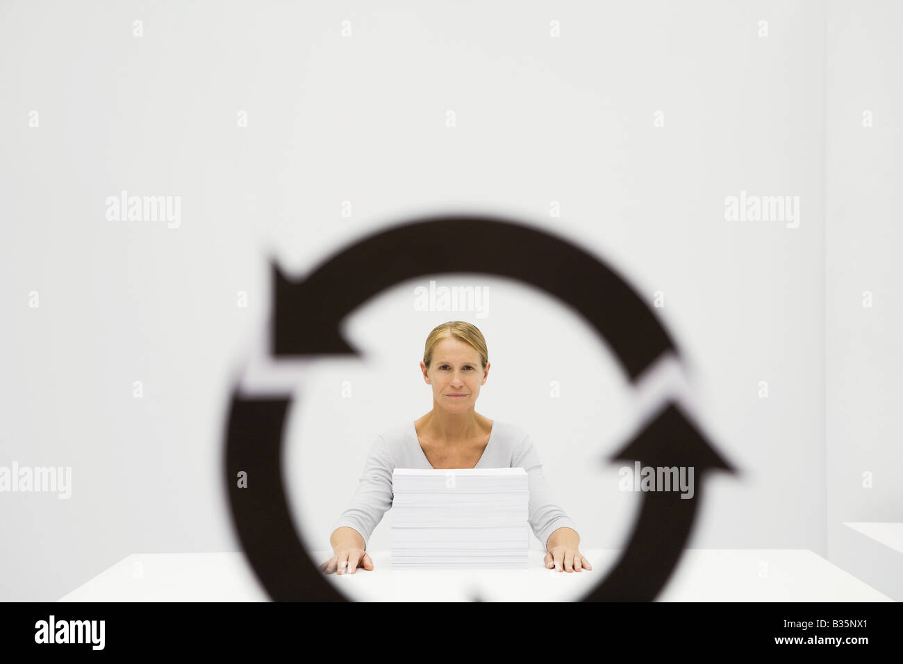 Professional woman sitting behind stack of paper, looking at camera through recycling symbol Stock Photo