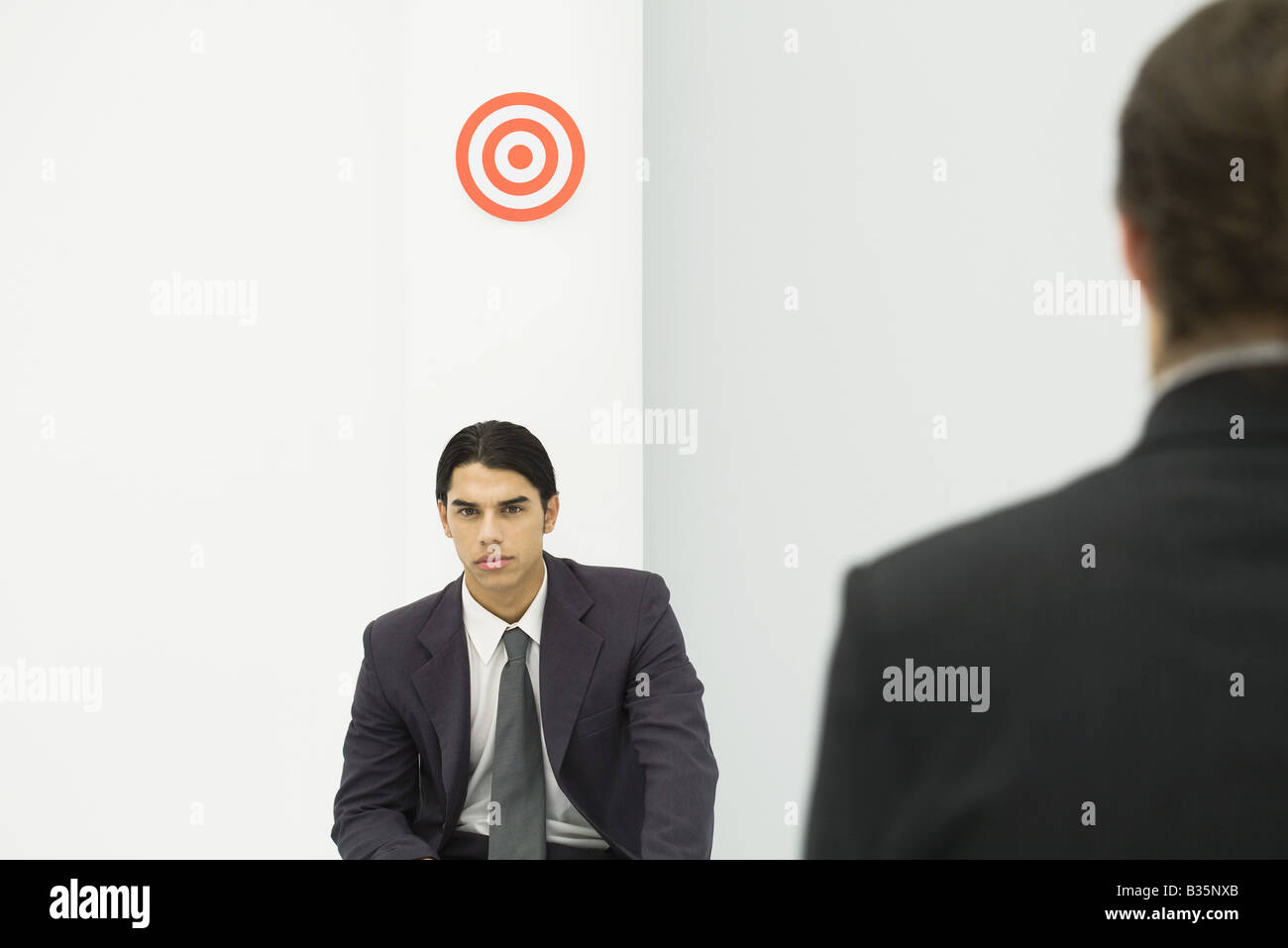 Young professional man sitting below bull's-eye, boss standing in foreground Stock Photo