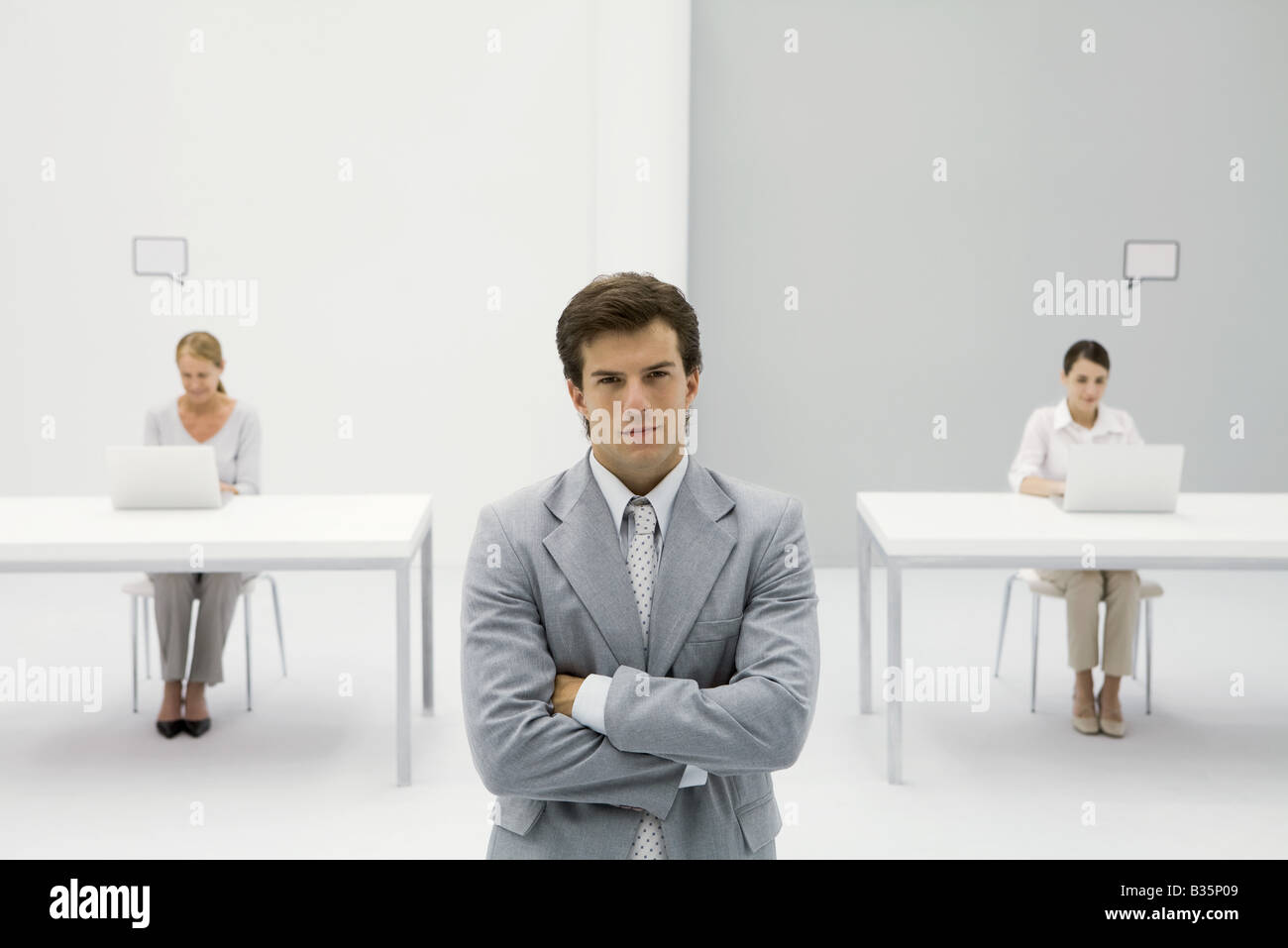 Businessman standing with arms crossed, smirking at camera, women working in background Stock Photo