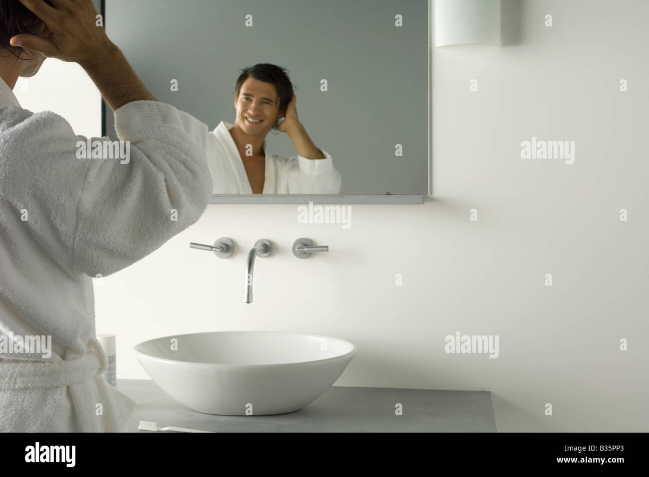 Man in bathrobe looking at self in bathroom mirror, hand in hair Stock Photo