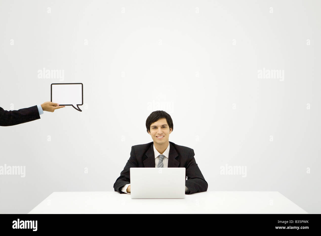 Young businessman sitting with laptop, smiling at camera, colleague holding up blank word bubble Stock Photo