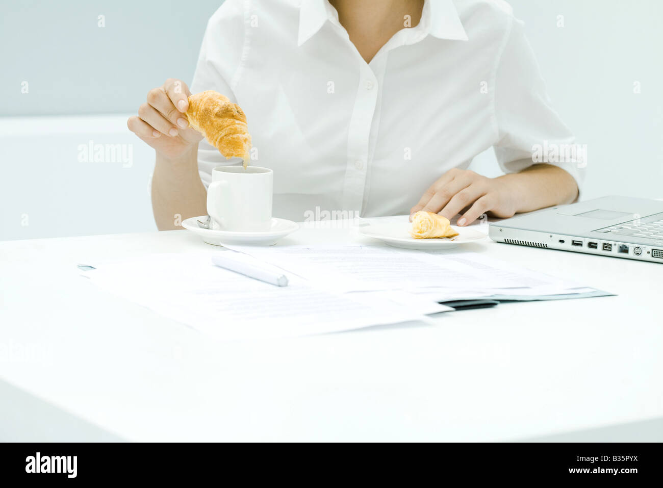 Woman dipping croissant into coffee, sitting at cluttered desk, cropped view Stock Photo
