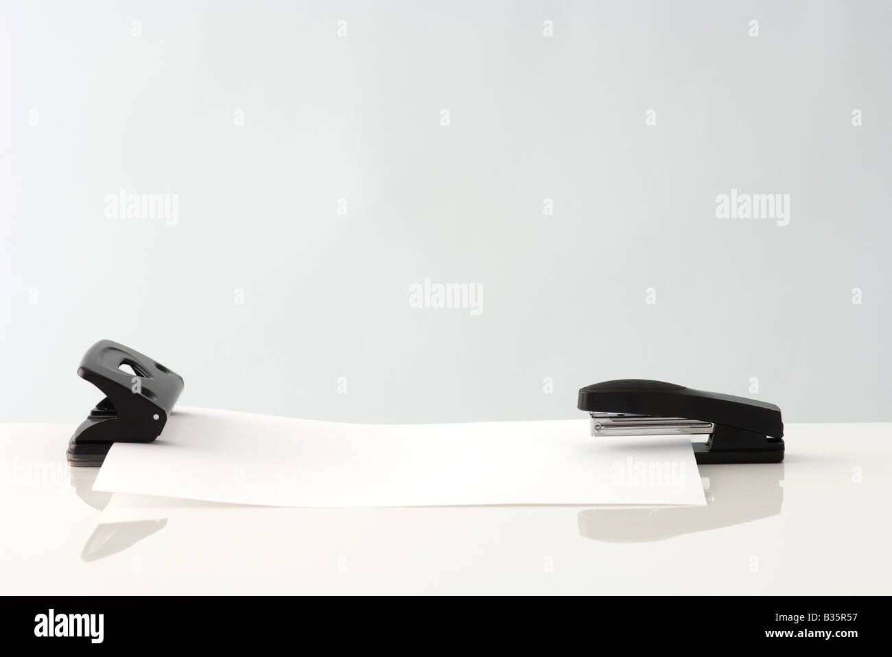 Sheet of paper, resting in hole puncher on one end and stapler on the other Stock Photo