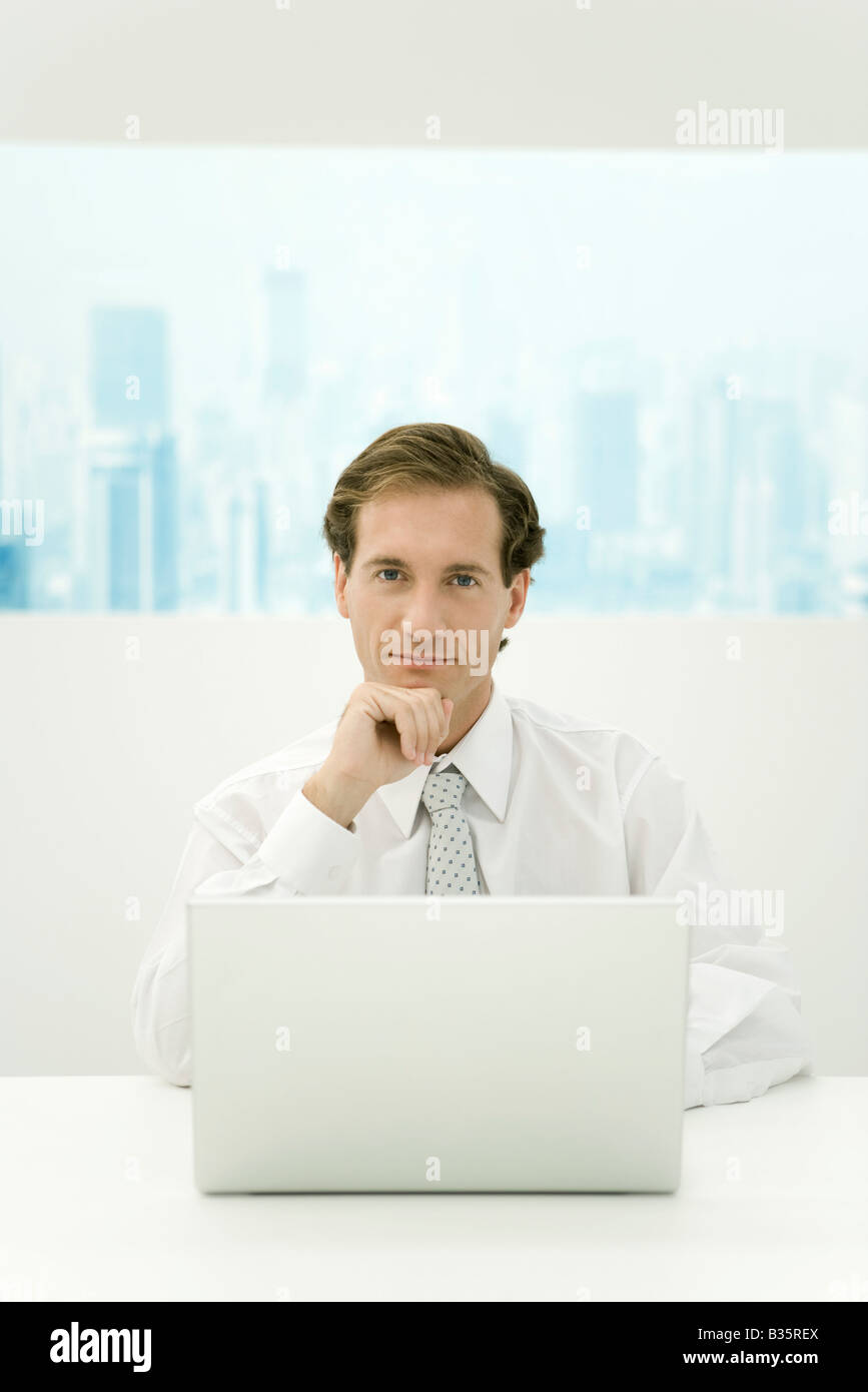 Businessman sitting with laptop computer, holding head, smiling at camera Stock Photo