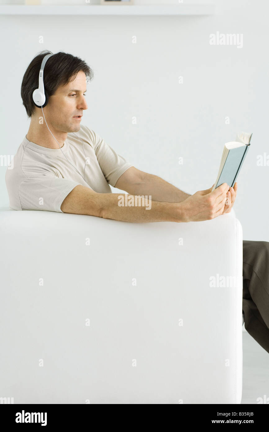 Mature man sitting in armchair, reading book and listening to headphones Stock Photo