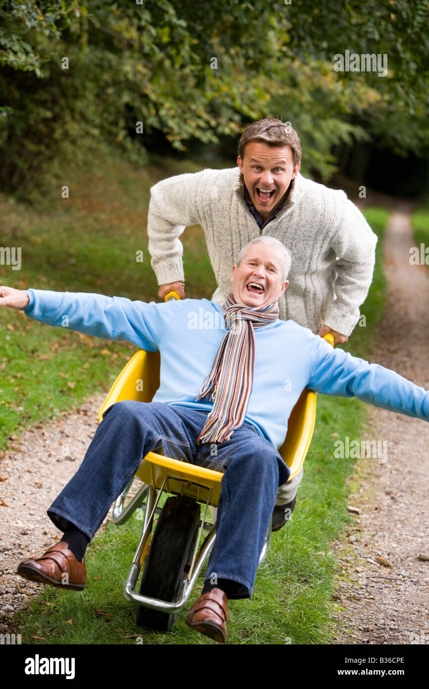 Man walking on path outdoors pushing other man in wheelbarrow and