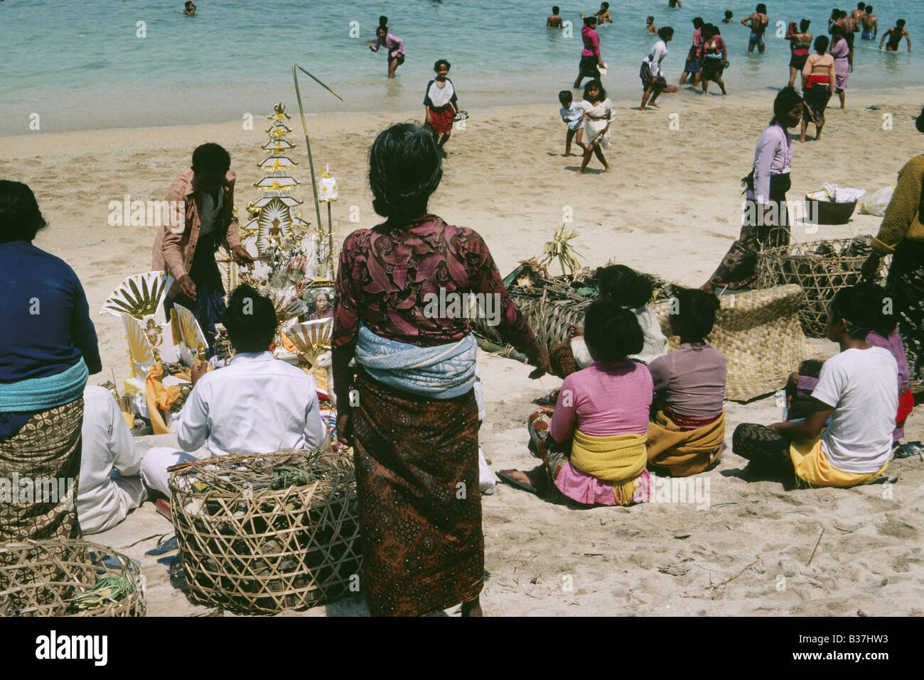 Ceremony held on beach Body in wicker bier People seated on sand Icons ornaments People in sea BEACH FUNERAL BALI INDONESIA Stock Photo