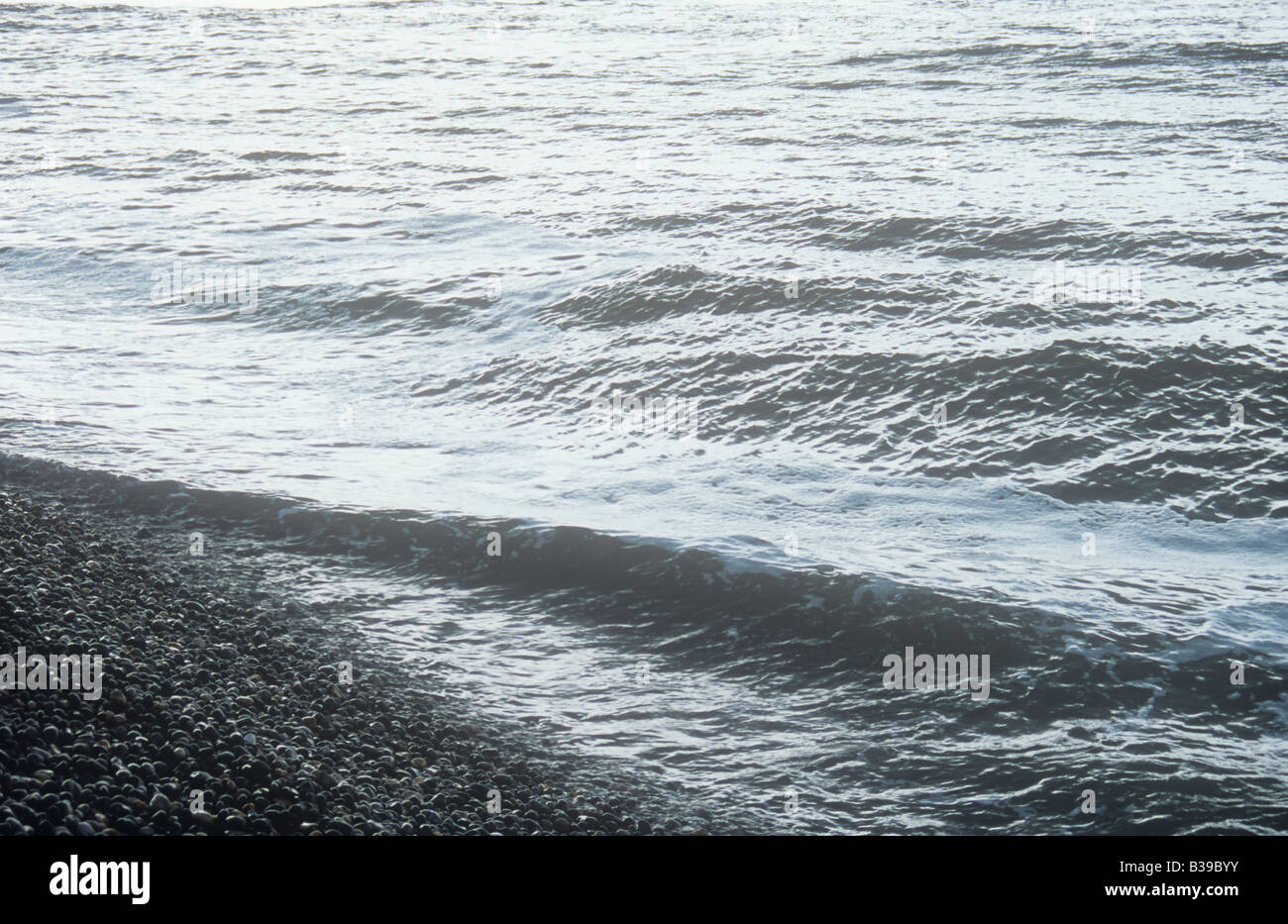 An uninviting silver grey sea with large swell breaking onto shore of smooth rounded gray pebbles Stock Photo