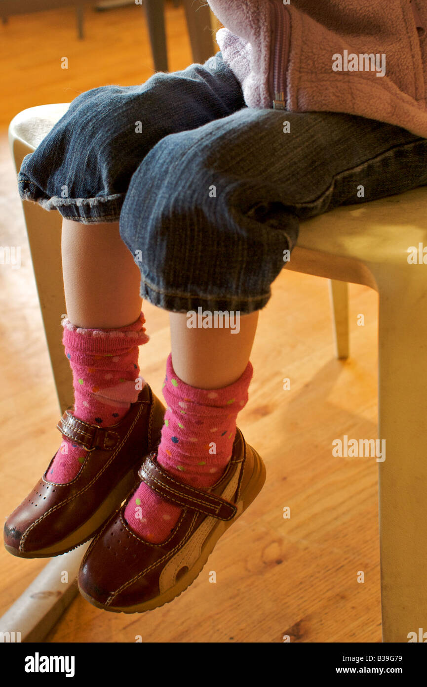 The legs and feet of 2-year-old girl as she sits on a chair in a cafe, wearing red mary jane shoes, pink socks and jeans. Stock Photo