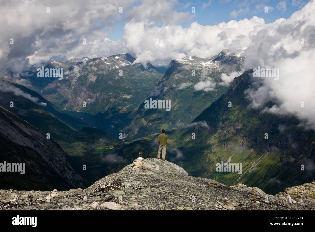 Aerial perspective 1 young adult man 20's looking out at Geiranger Fjord, Norway from Dalsnibba mountain plateau, dramatic blue sky, fluffy clouds Stock Photo