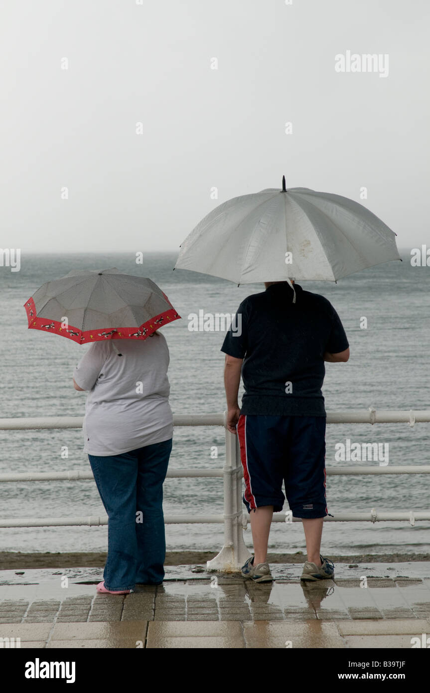 two 2 people on summer holiday vacation standing in the rain under umbrellas on Aberystwyth seaside promenade August 2008 Stock Photo