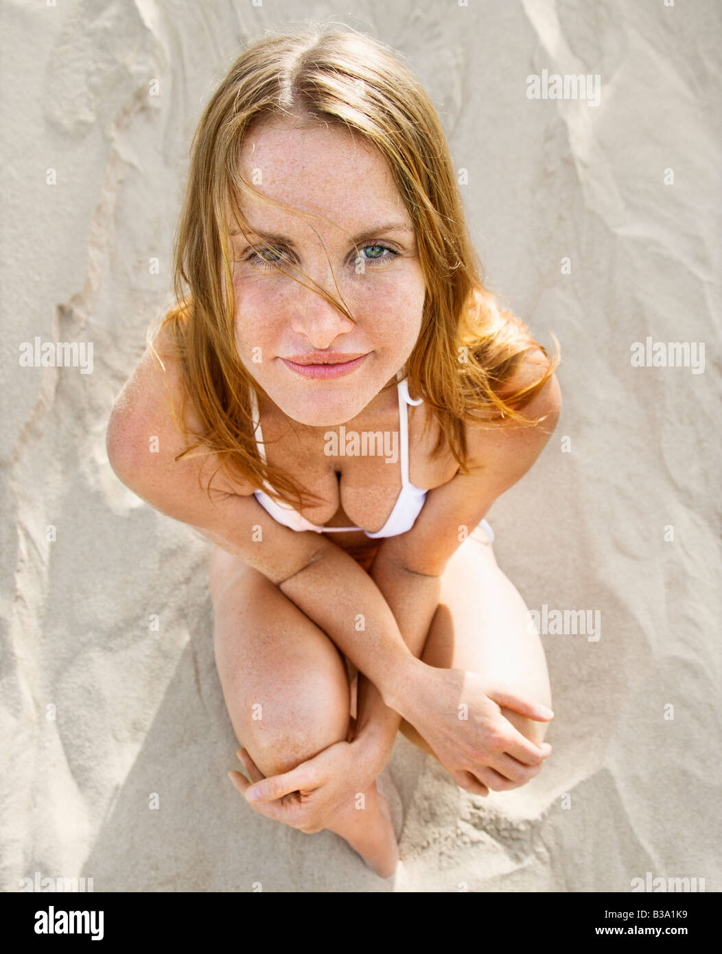 High angle view of redheaded woman sitting on beach wearing bikini looking at viewer Stock Photo