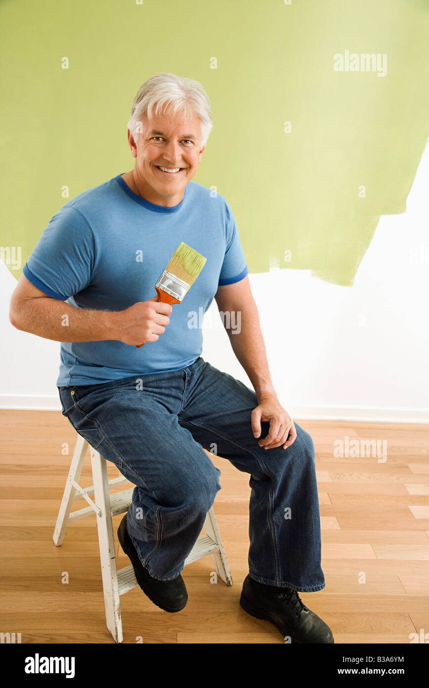 Portrait of smiling adult man sitting in front of half painted wall with paintbrush Stock Photo