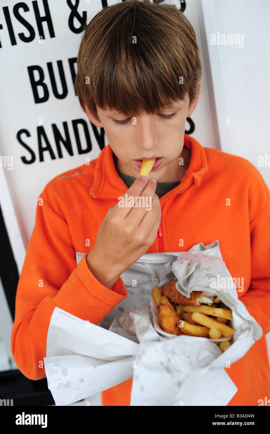 A young boy eating fish and chips out of newspaper outside a fish and chip take away restaurant in Lyme Regis, Dorset, UK Stock Photo
