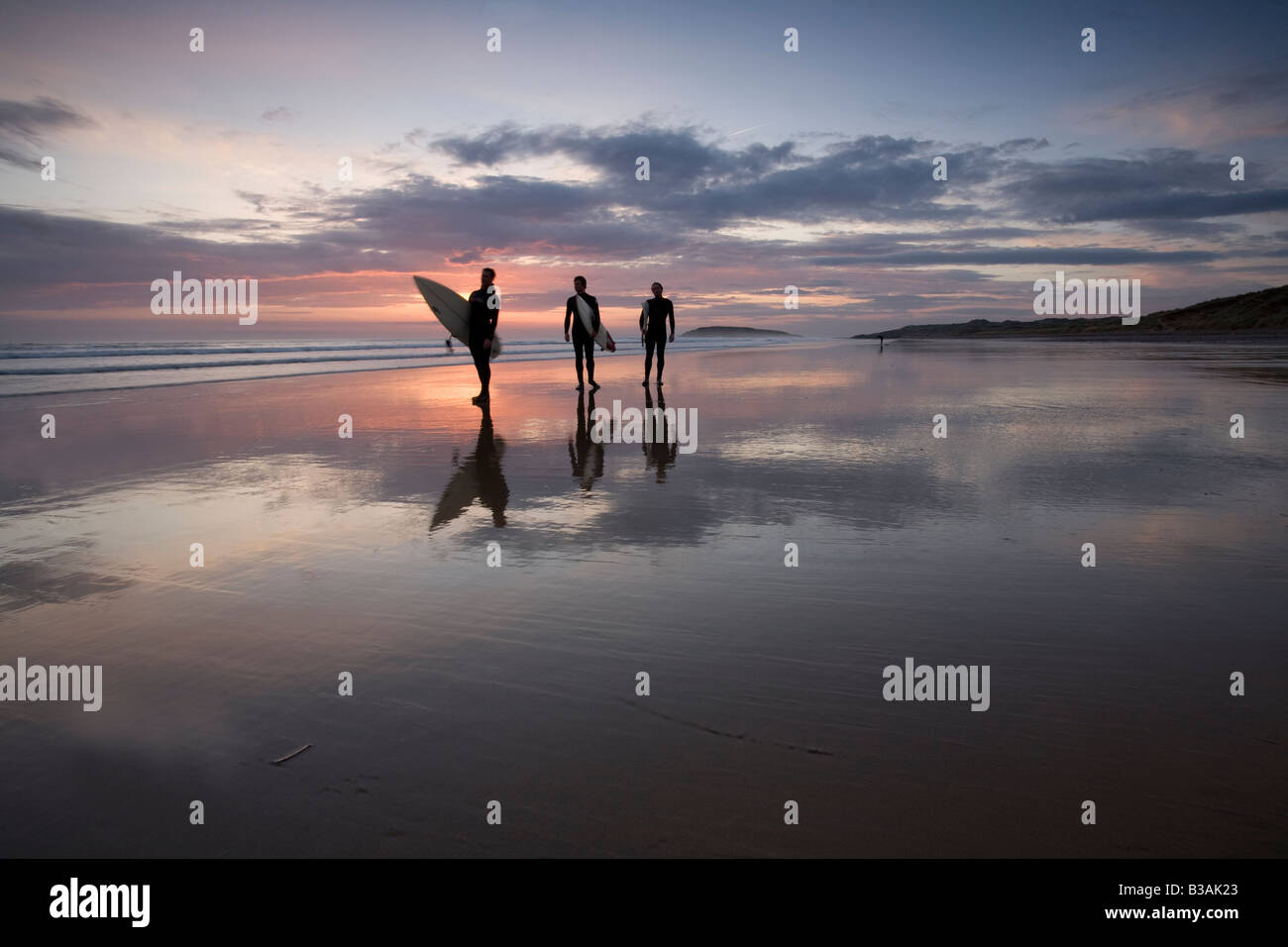 Three people, surfers, walking towards camera in silhouette on beach at sunset with reflections of sunset and clouds in the sand Stock Photo