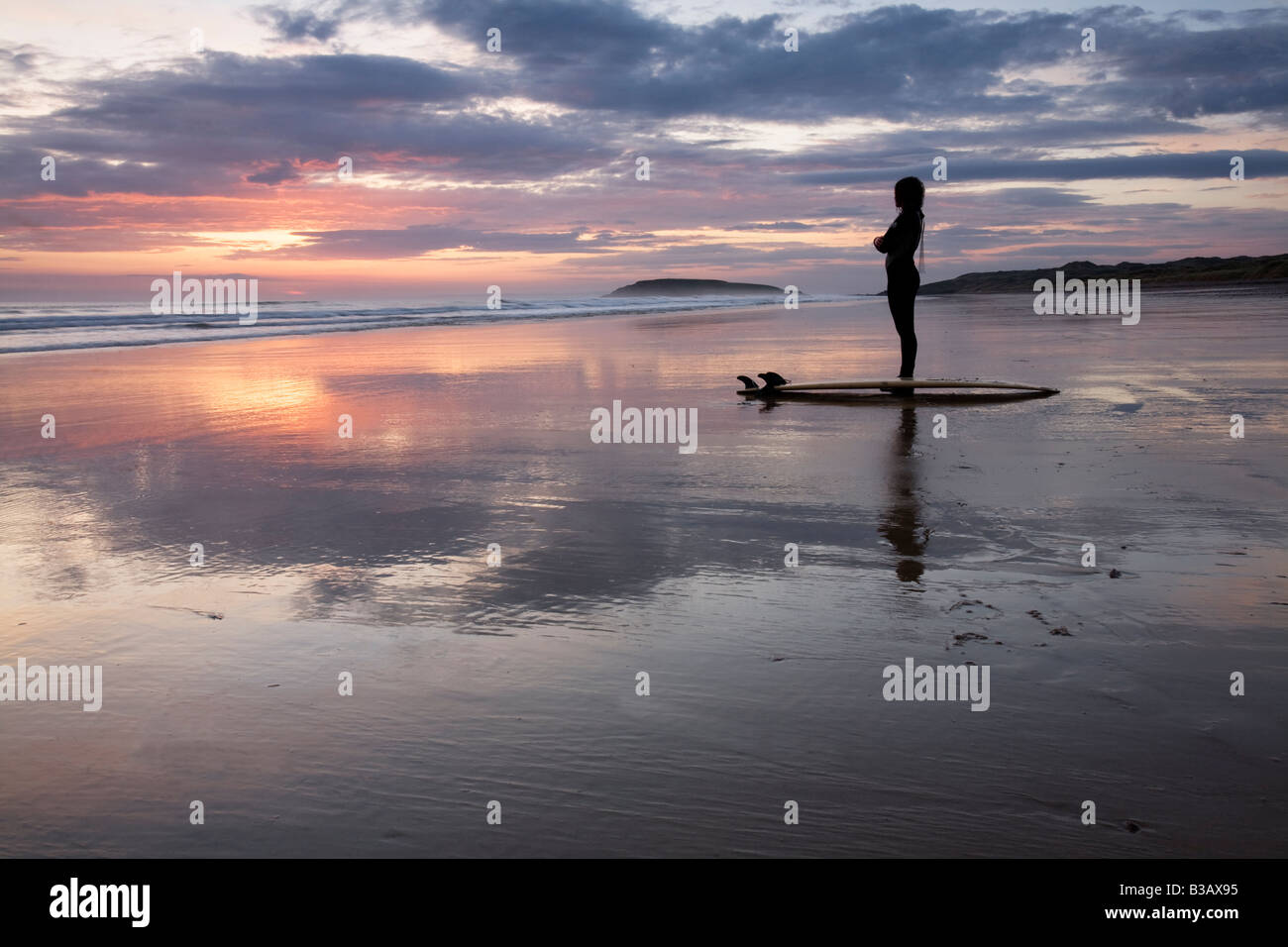 One woman surfer standing in silhouette on beach at sunset with reflections of sunset and clouds in the sand Stock Photo