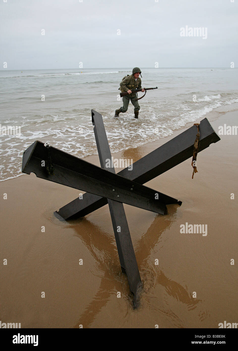 An actor dressed as an D-Day  American soldier runs up Omaha Beach Normandy France passed a 'Hedgehog' tank trap. Stock Photo