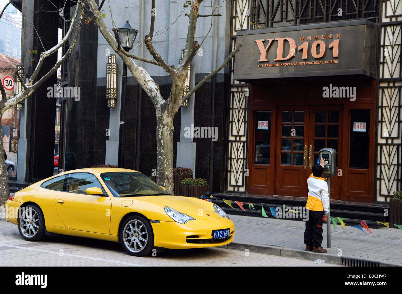 China, Shanghai. French Concession area - a young boy putting coins in the parking meter next to a yellow Porshe. Stock Photo
