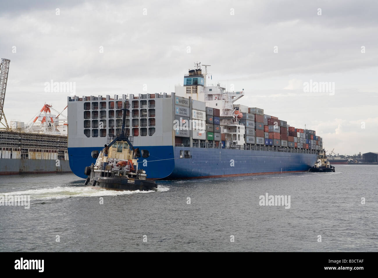 Container ship, the Maersk Dampier, is guided into the port of Fremantle, Western Australia, by two hard working tug boats Stock Photo