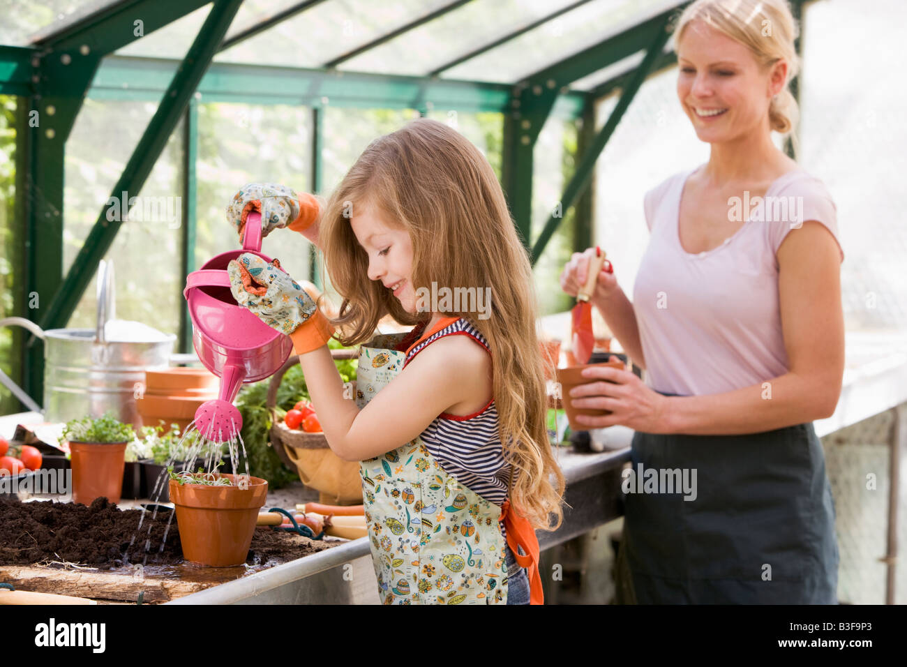 Young girl in greenhouse watering plant with woman holding pot smiling ...