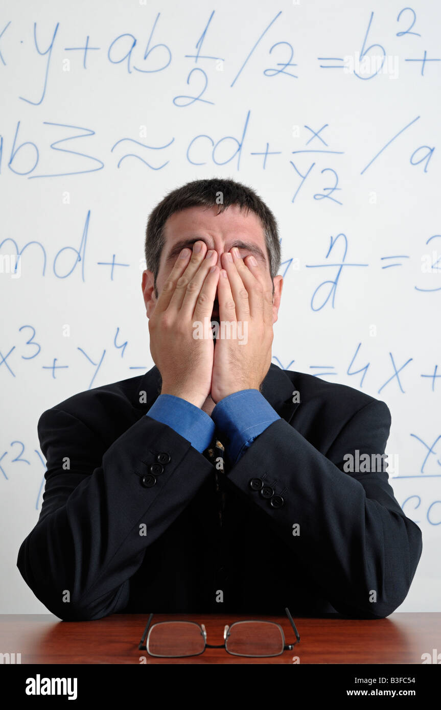 Tired Business Man Rubbing His Eyes Whilst Sitting at His Desk in Front of a Whiteboard Full of Calculations Stock Photo