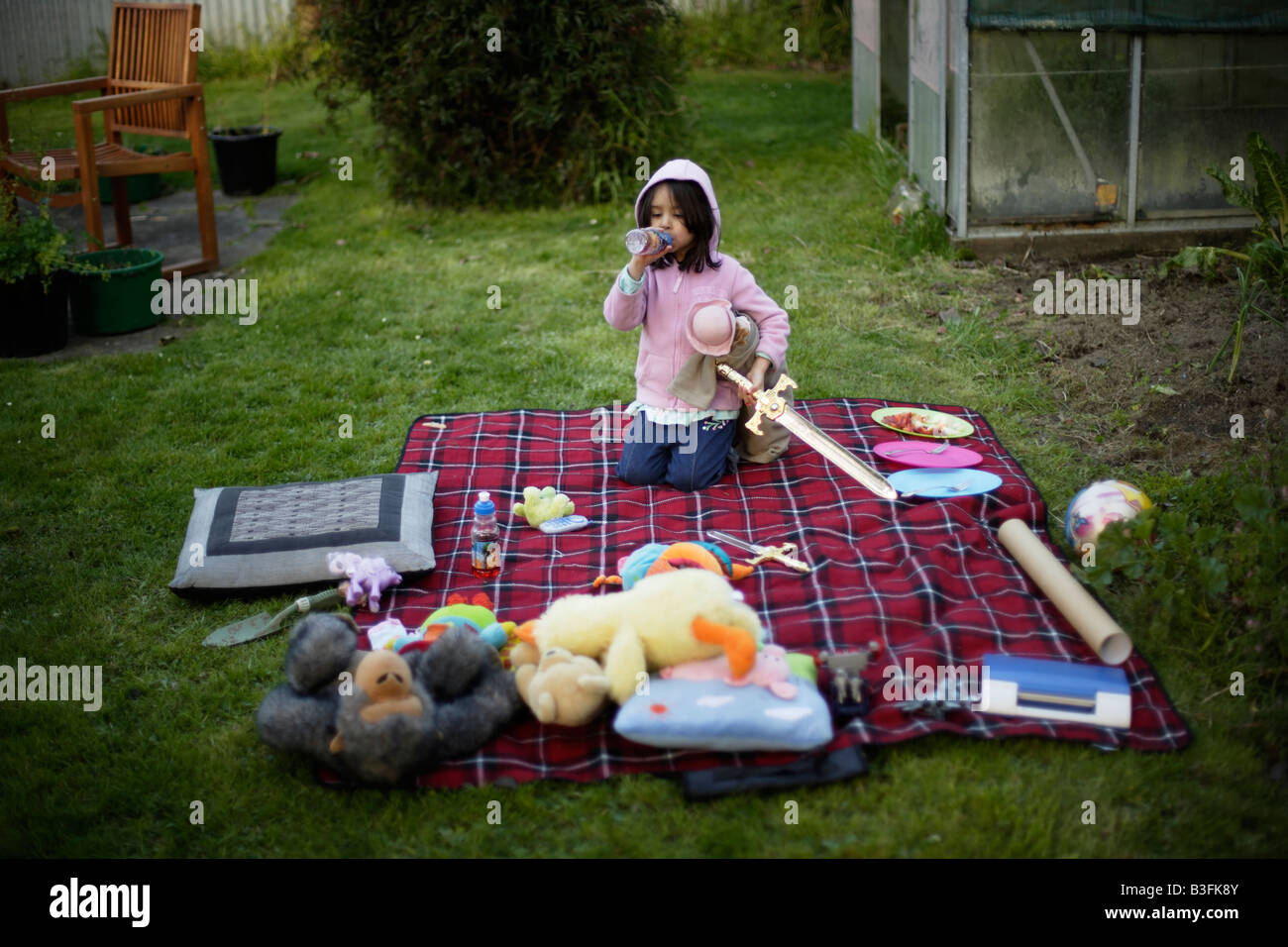 Picnic in the garden Five year old girl drinks juice from a bottle amongst soft toys laid out on picnic blanket on lawn Stock Photo
