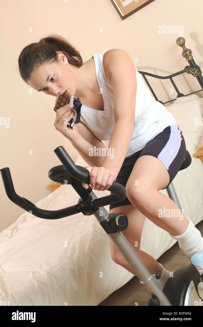 a girl fitness training on her exercise bike looking guilty whilst eating a chocolate bar Stock Photo