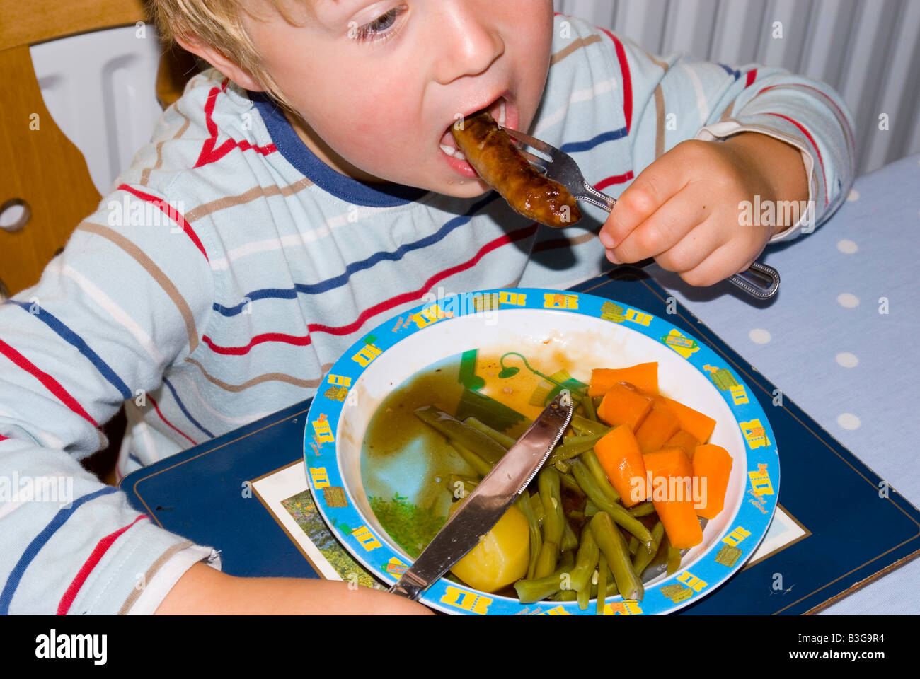 Four year old boy eating a healthy dinner with sausages, fresh vegetables and gravy. Stock Photo