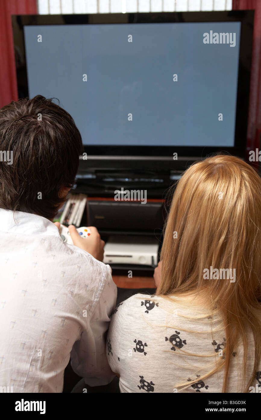 young late teenage couple sitting together playing a video game on a large screen plasma tv in a living room Stock Photo
