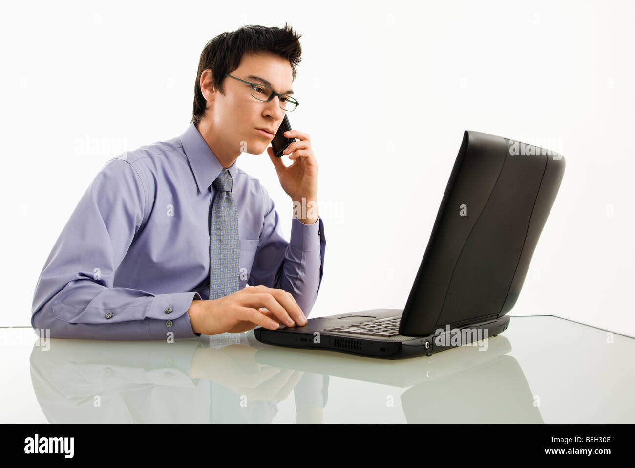 Asian businessman sitting at desk working on laptop talking on cellphone Stock Photo