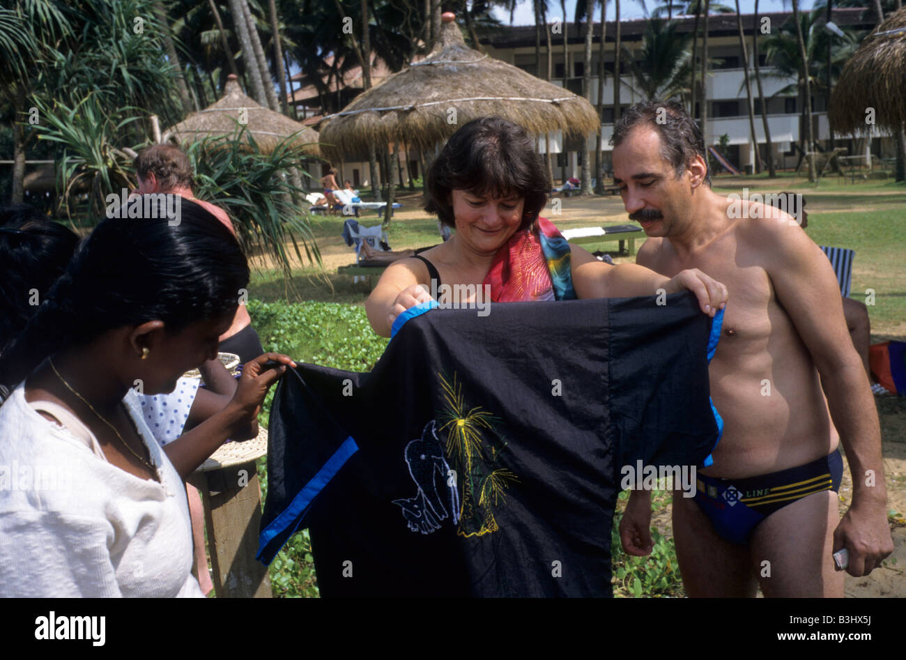 A western couple at a beach resort look to buy textiles from a Sri Lankan woman on the southern coast in Mathura, Sri Lanka. Stock Photo
