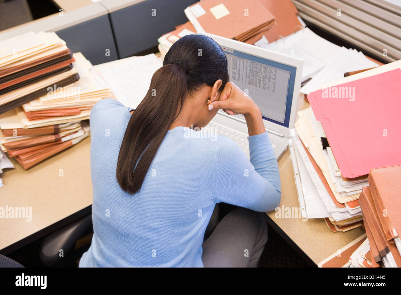 Businesswoman in cubicle with laptop and stacks of files Stock Photo
