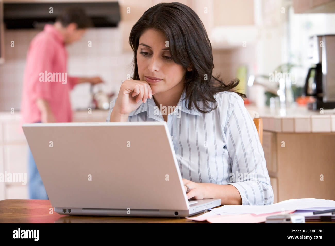 Woman in kitchen with paperwork using laptop with man in background Stock Photo