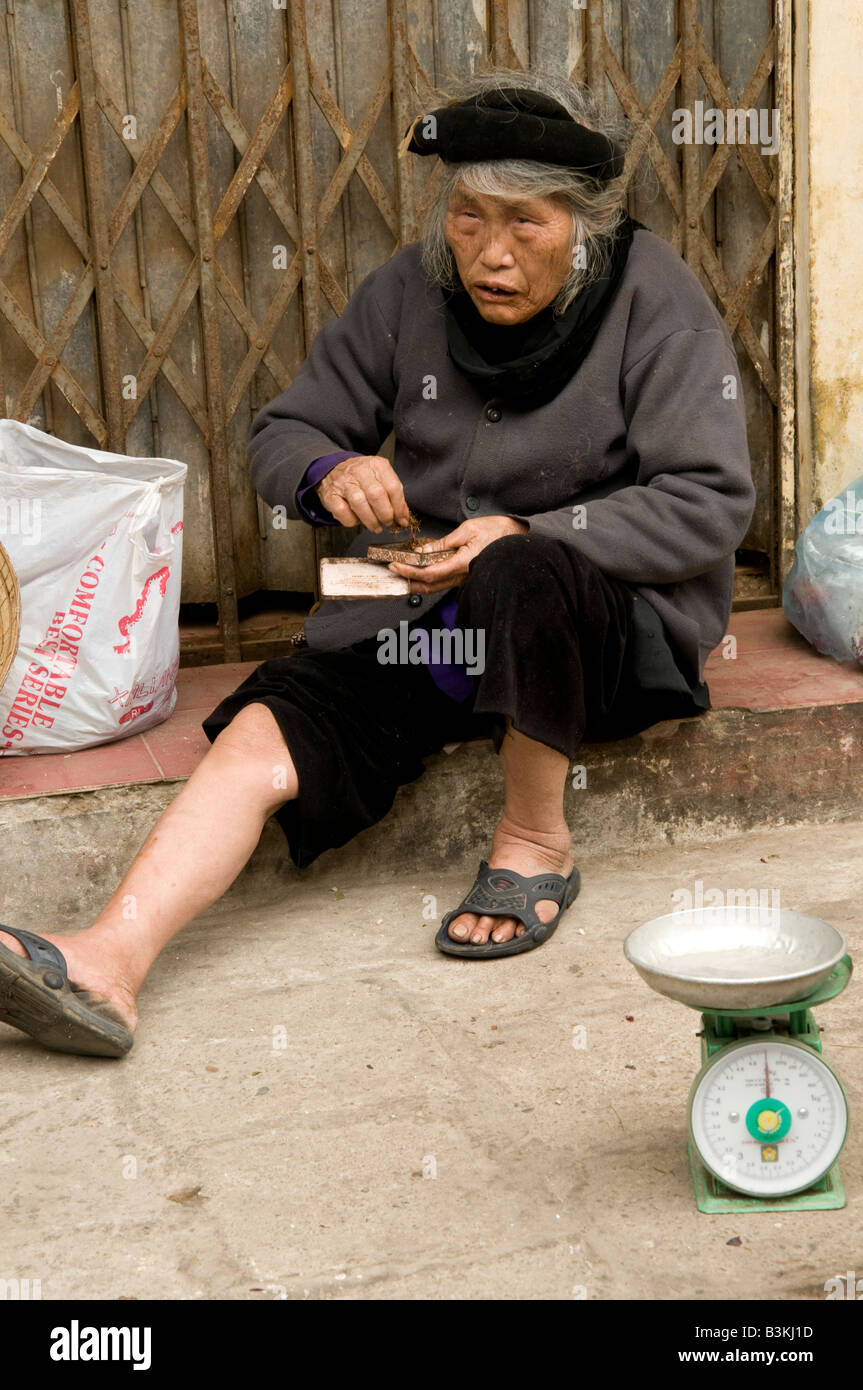 An elderly woman rolls herself a cigarette while sitting on a shop doorway in Hanoi Vietnam Stock Photo