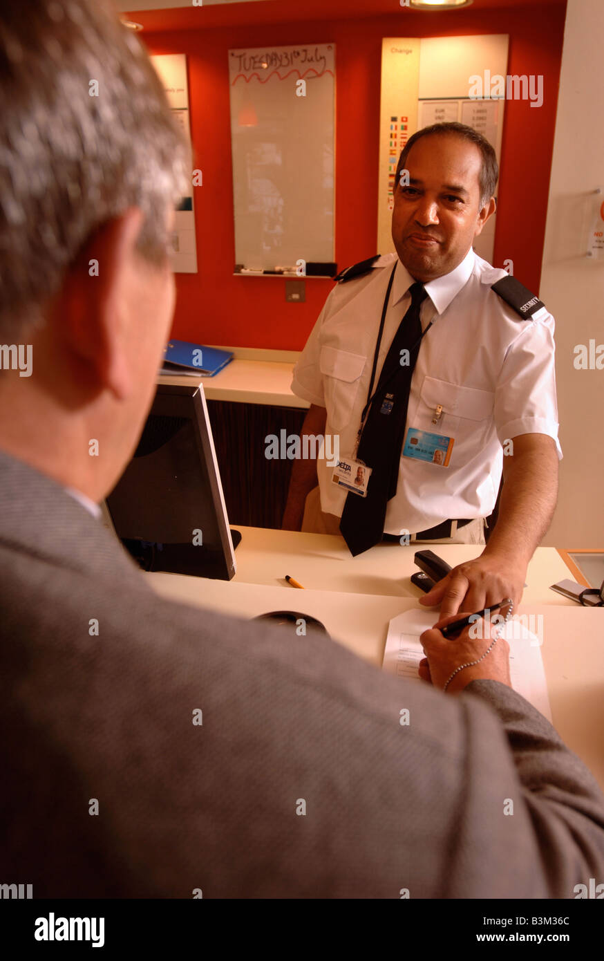 A GUEST SIGNS IN HELPED BY A SECURITY GUARD AT A HOTEL RECEPTION DESK