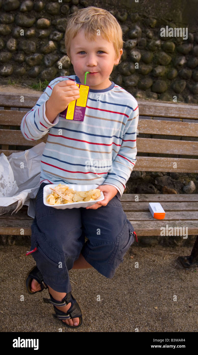 Five year old boy eating chips outside sitting on bench and having a drink Stock Photo