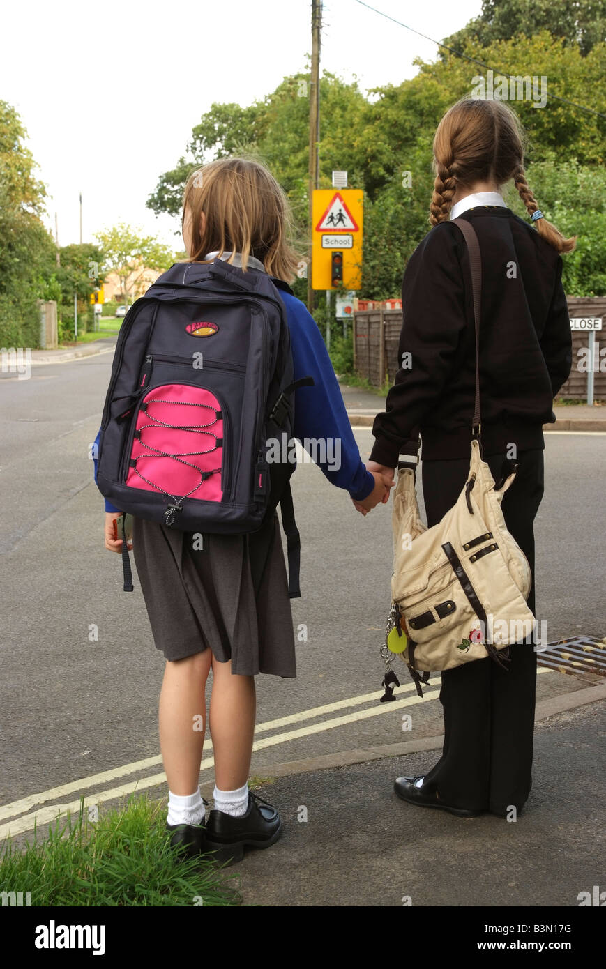 Schoolgirls in uniform wait to cross a road on the school route England UK Road safety wait at the kerb before crossing a road Stock Photo
