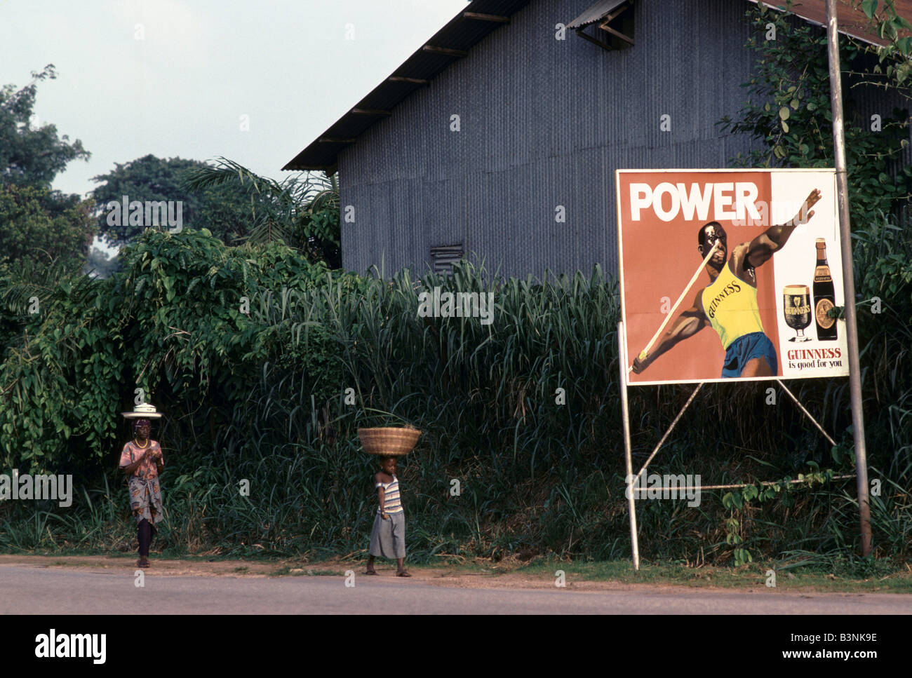 CHILDREN CARRYING ITEMS ON THEIR HEADS, WALKING ALONG RURAL ROAD, PASSING BILLBOARD ADVERTISING POSTER FOR GUINNESS Stock Photo
