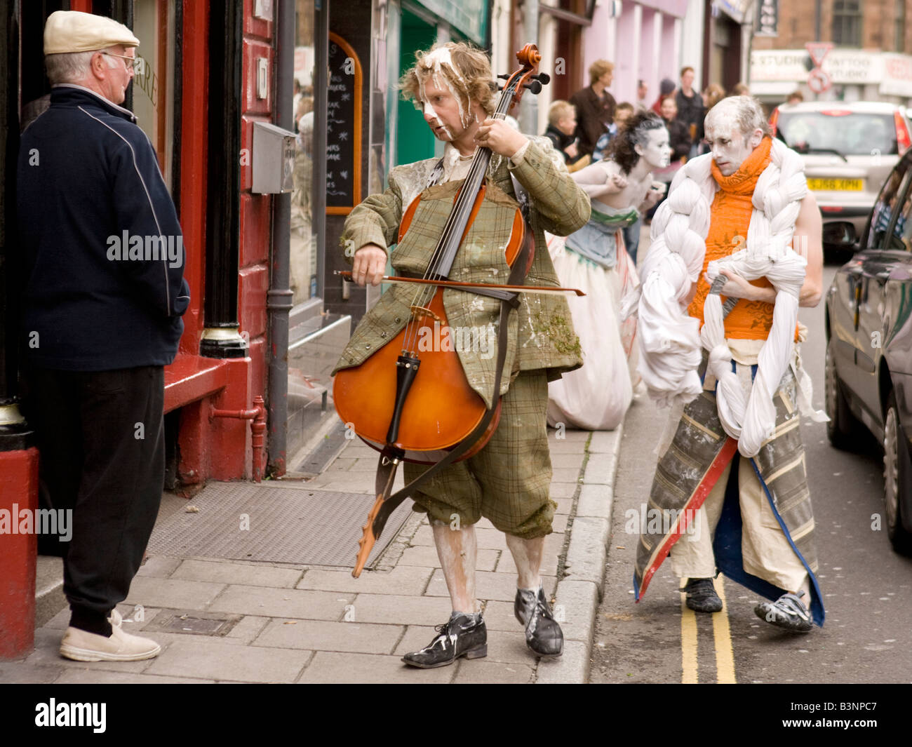 Street theatre strange costume musician playing cello outside pub old man with cap part of Gaelforce Art Festival Dumfries UK Stock Photo