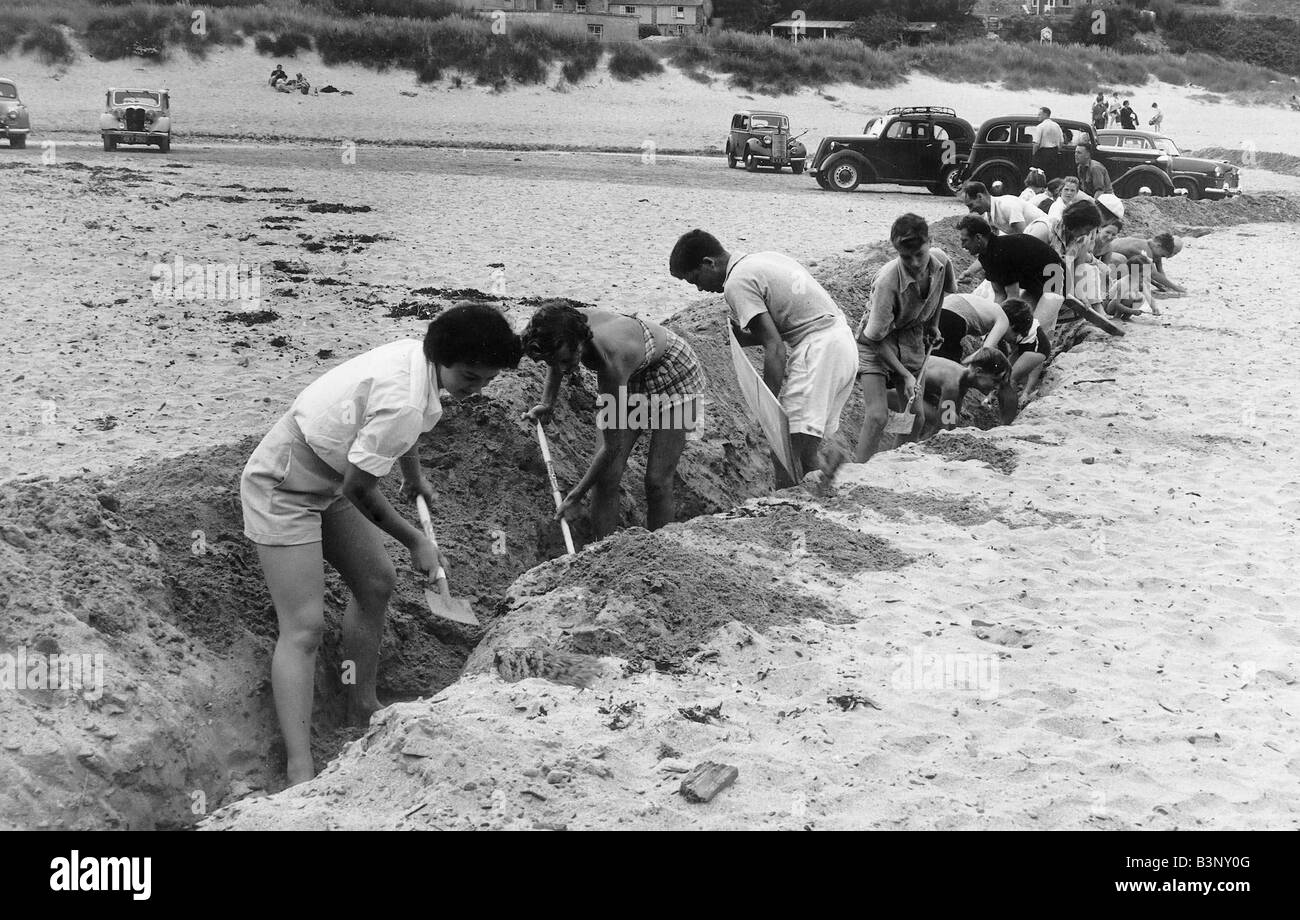 Porthcothan Bay Cornwall 1950 s Holiday Makers dig a trech in the sand to stop cars from being driven on to the beach The trench was 150 yards long and 2 feet deep once it was completed sea water was diverted into it to stop the cars One bridge wide enough for prams was put across the trench cars in the backgroung are prevented from driving on to the beach Stock Photo