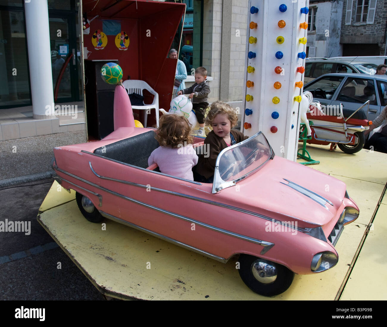 Barneville, Normandy, France. Two little girls in a pink model Cadillac on a merry go round (carousel) in the weekly market Stock Photo