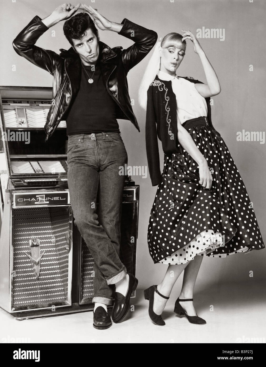 Models posing wearing fifties style fashion man wearing jeans black t shirt  and black leather jacket combing his hair standing next to a girl wearing a  wide floppy skirt and beaded cardigan
