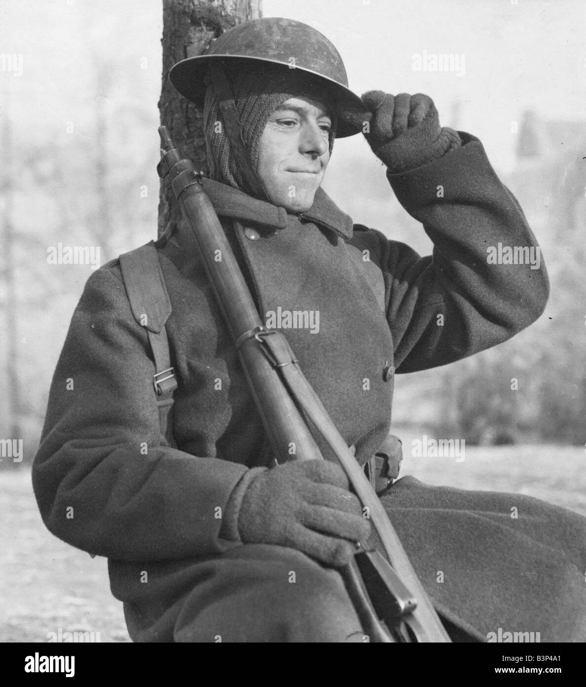 Driver David Smith of the Royal Engineers December 1944 in Belgium thinking of home Sitting up against tree holding his rifle and adjusting his tin helmet Wearing thick overcoat and gloves Stock Photo