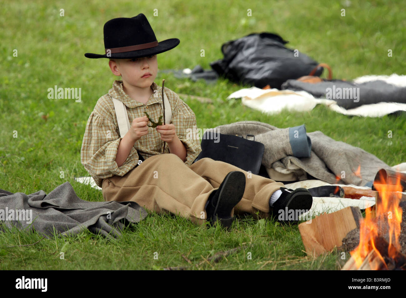 A boy in an 1850s outfit playing with a leaf by a campfire at a Civil War Encampment Reenactment Stock Photo