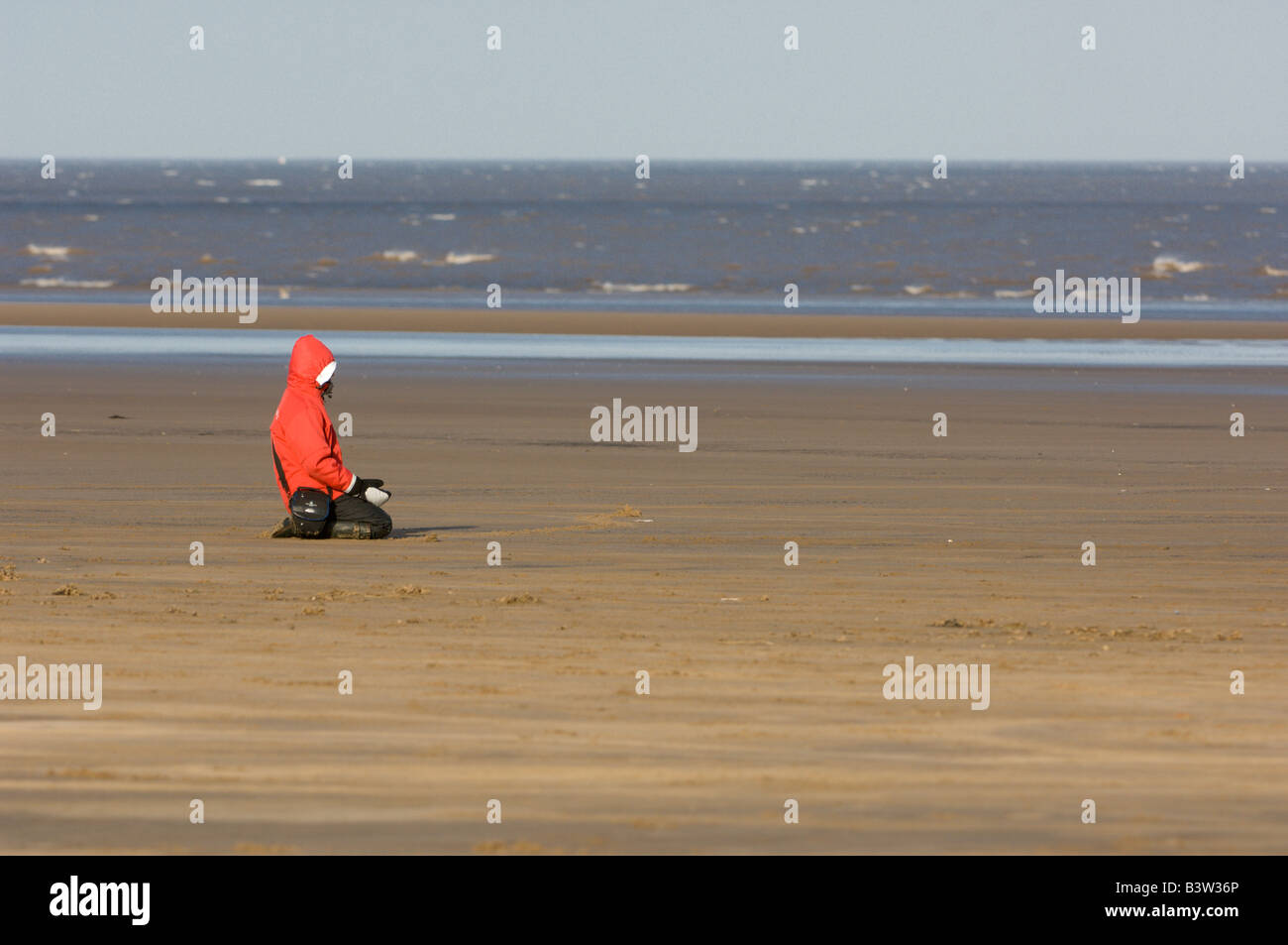 Young person sitting on the beach wearing a red coat, Donna Nook, UK Stock Photo
