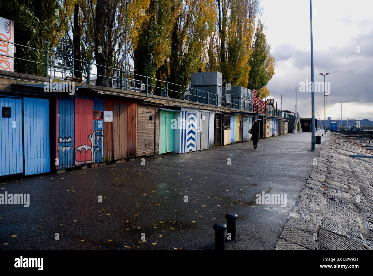 Blustery fall day along the port of Neuchatel.  A walker strolly past clorfully painted fishing sheds. Stock Photo