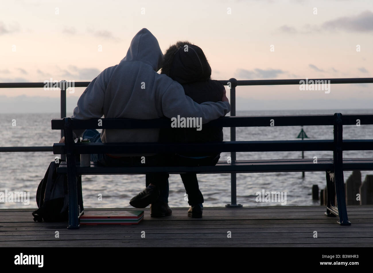 Young teenage couple wearing hoodies sitting and cuddling at sunset, Aberystwyth Wales UK, summer evening, seaside Stock Photo