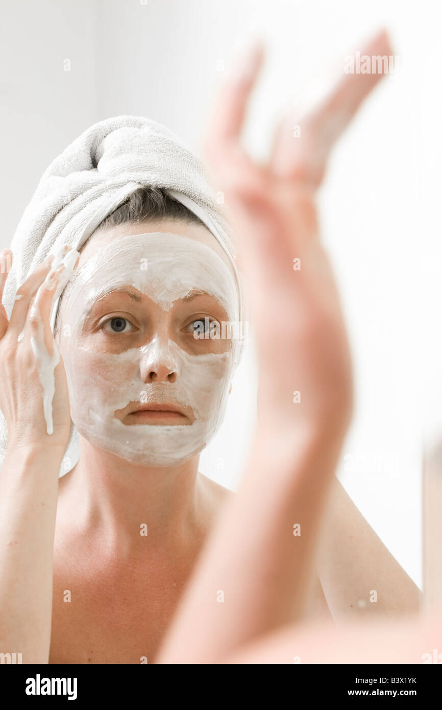 Woman applying a face mask whilst looking in a mirror, wearing a towel on her head. Vertical Shot Stock Photo